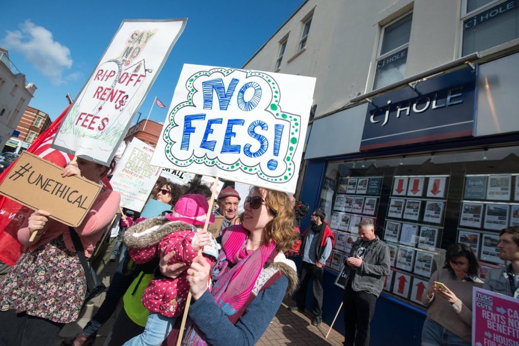 Protestors holding up signs that say "No Fees!", "Say no to rip off rents + fees" and "#unethical".